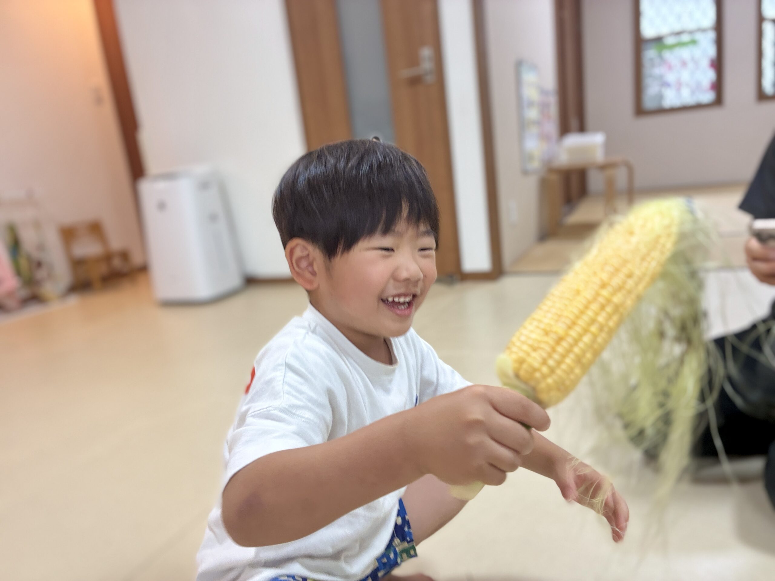 トウモロコシの皮むきを楽しみ、黄色い実を見つけて笑顔を見せるこども