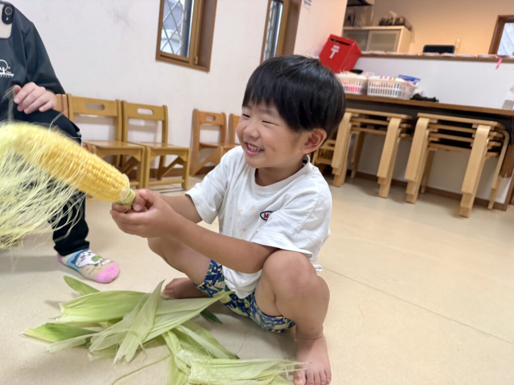 トウモロコシの皮むきを楽しみ、黄色い実を見つけて笑顔を見せるこども