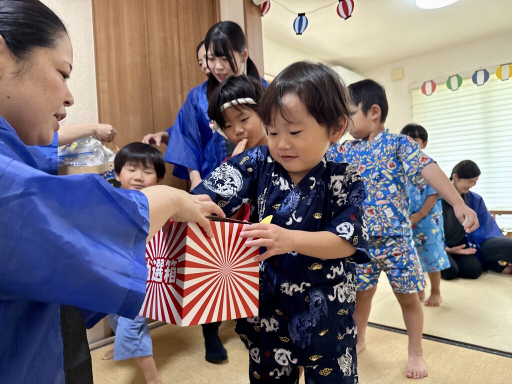 赤白の抽選箱に手を入れる甚平姿の子どもと箱を支える大人