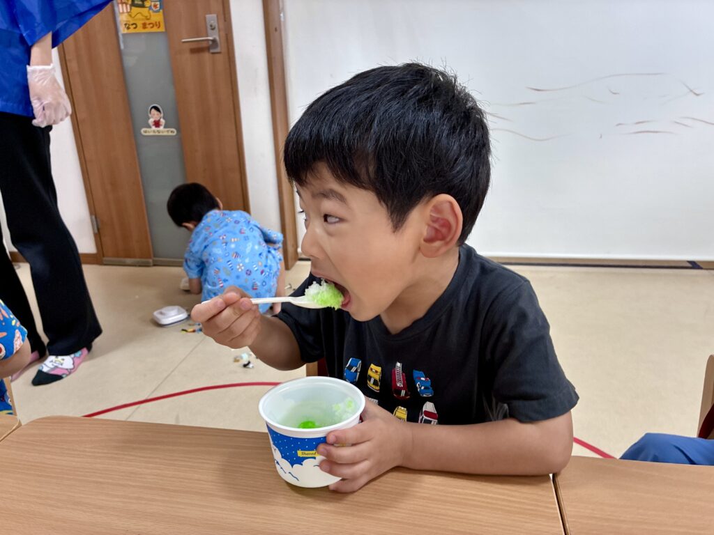 テーブルでカップを持ち、スプーンで緑色のかき氷を食べる子ども