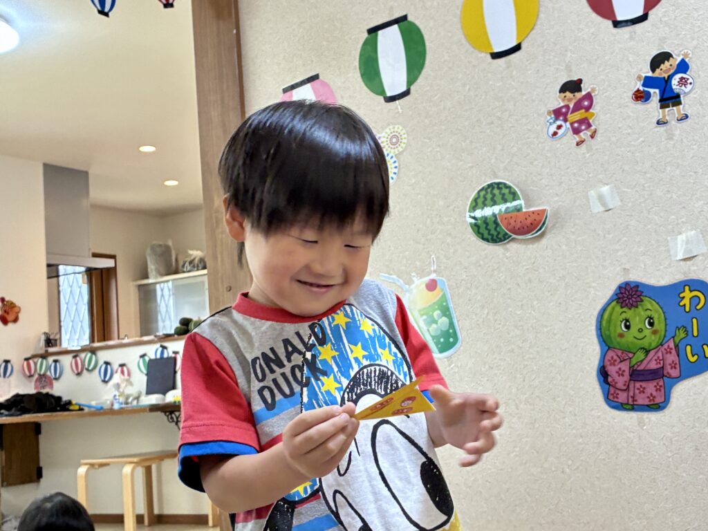 夏祭りの飾りの前で小さな黄色いカードを手に持つ子ども