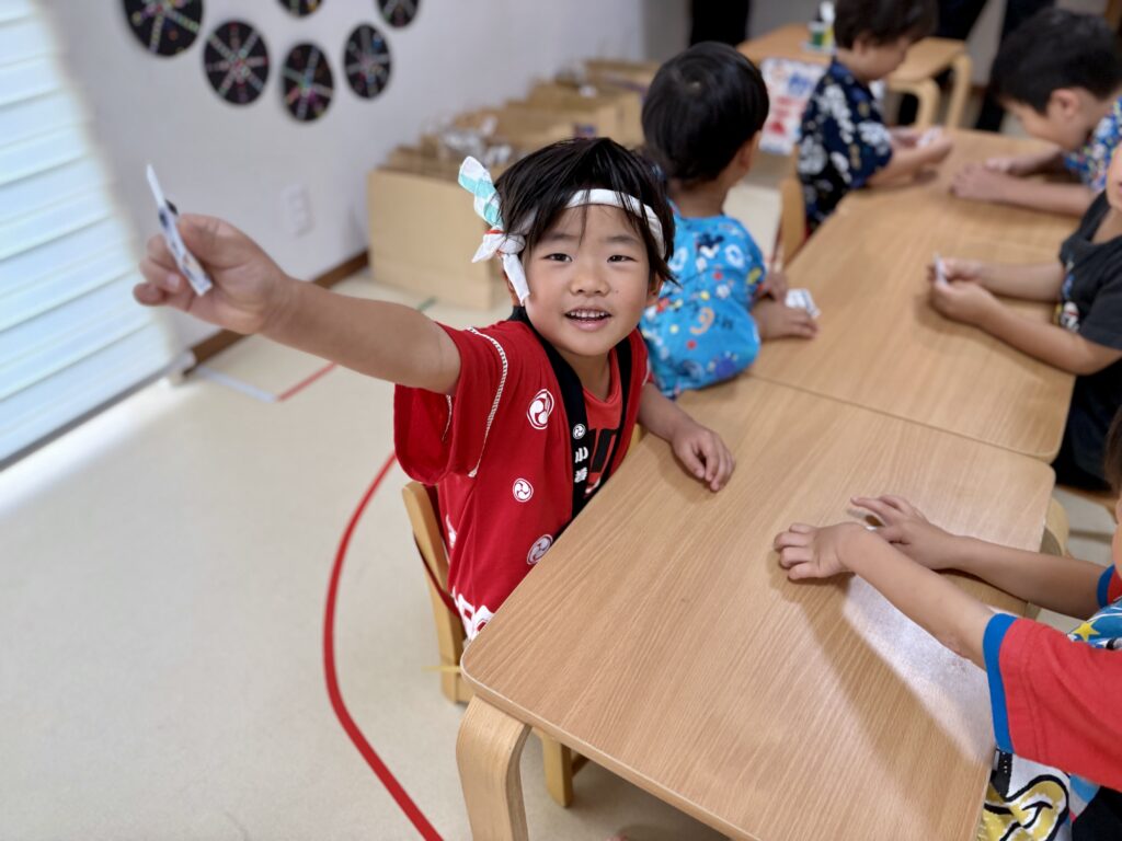 赤い法被と鉢巻きを着け、テーブルで小さな紙を手に掲げる子ども