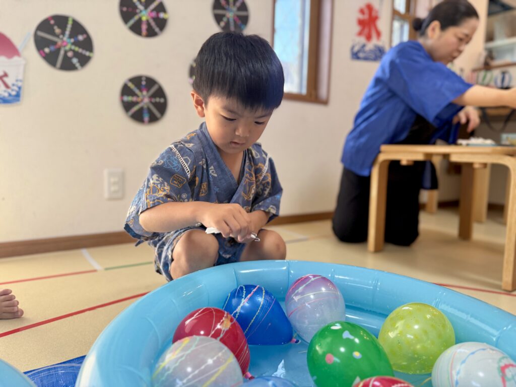 水ヨーヨーの入った青い水槽の前で釣り針を持つ浴衣姿の子ども