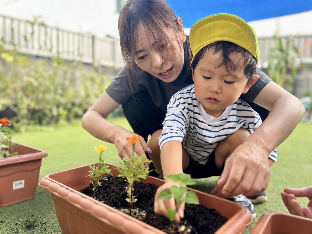 スタッフと一緒にプランターの花の苗へ土をかけるこども
