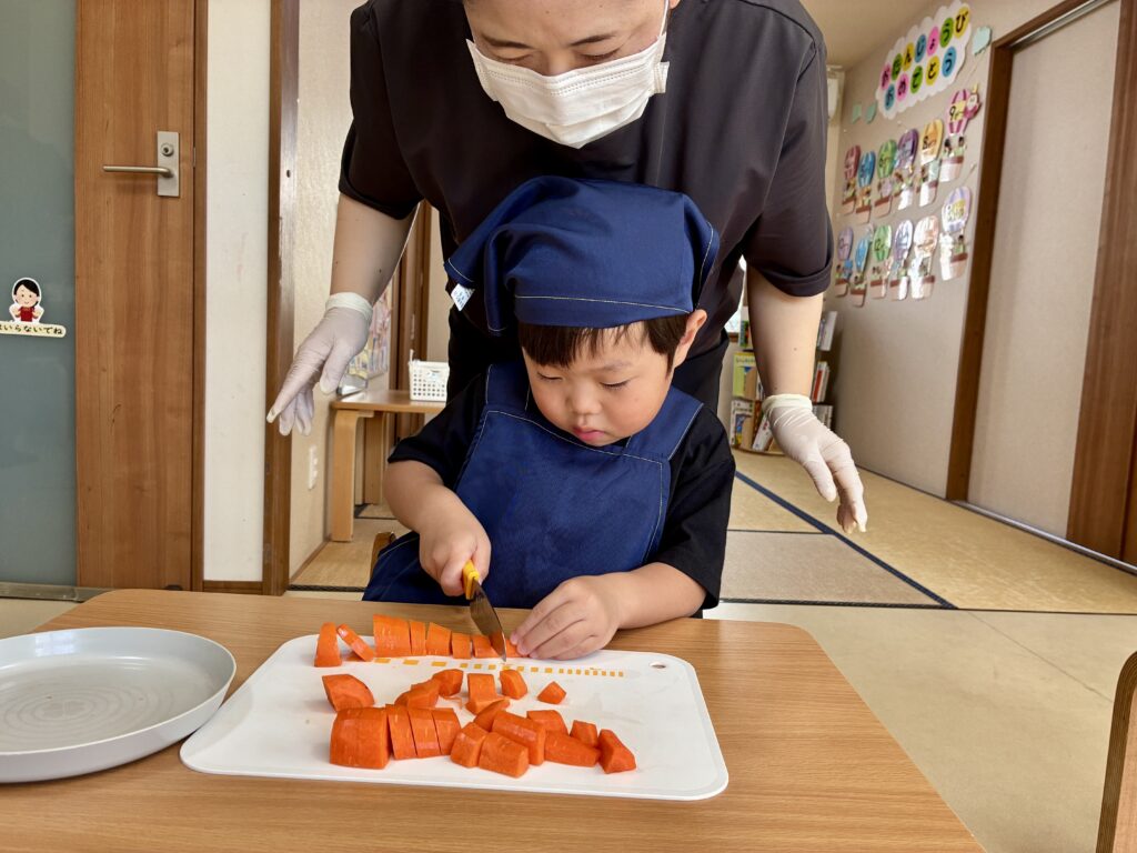 エプロンと三角巾姿でにんじんを包丁で切るこども。後ろでスタッフが見守る