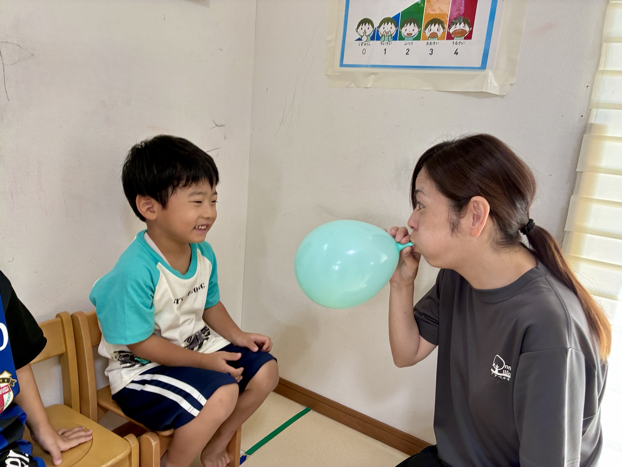 職員が風船をふくらませる様子を、となりで見て笑うお友達