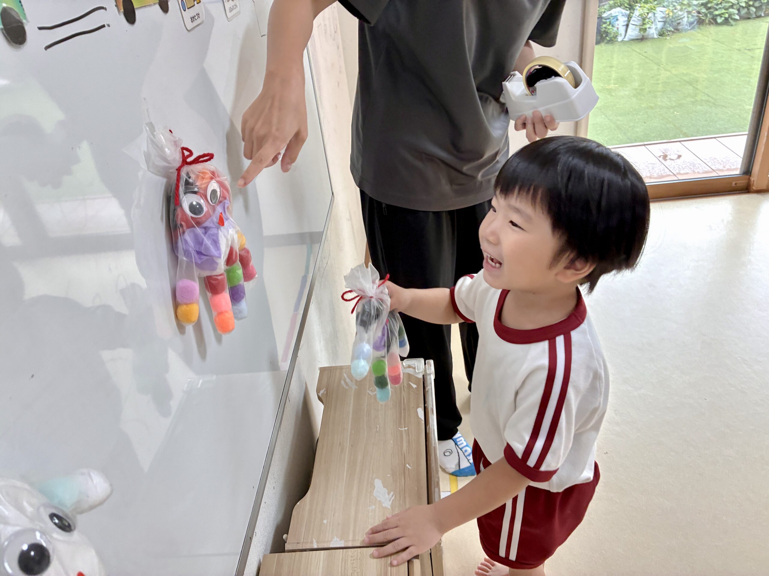 ホワイトボードに飾ったおばけの前で、自分のおばけを持って笑顔の子どもと職員