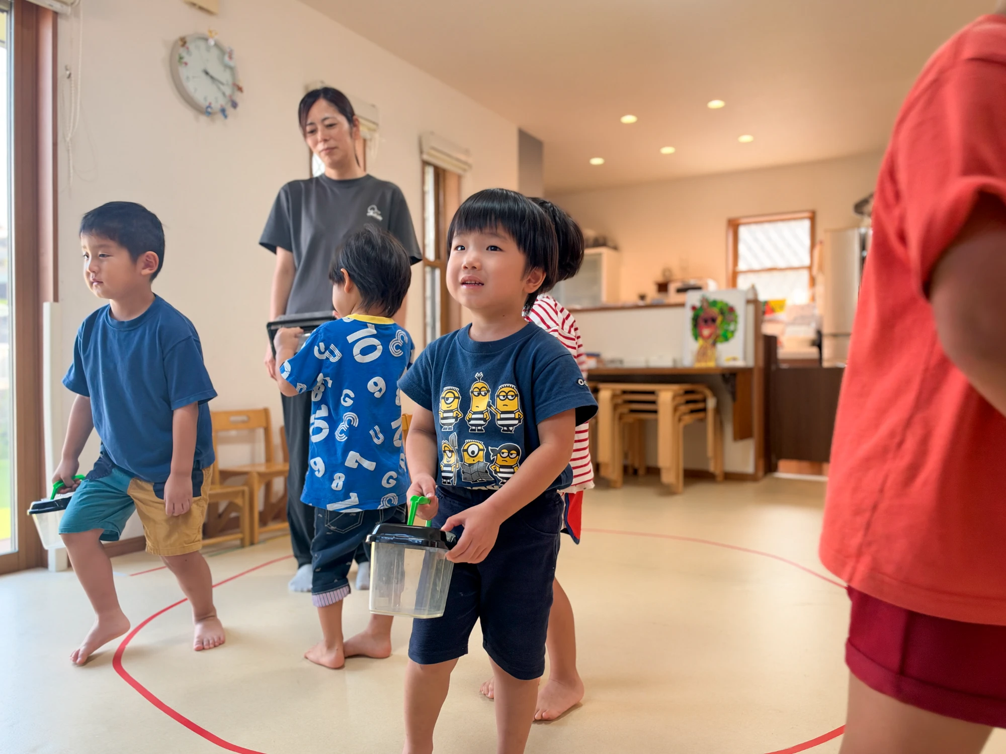 虫かごを持って室内を歩く子どもたちと見守る職員